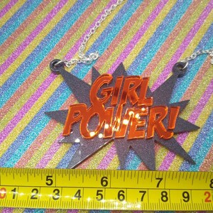 May include: A silver chain necklace with a pendant that reads "GIRL POWER!" in orange letters. The pendant is in a black starburst shape. A yellow measuring tape is at the bottom of the image.