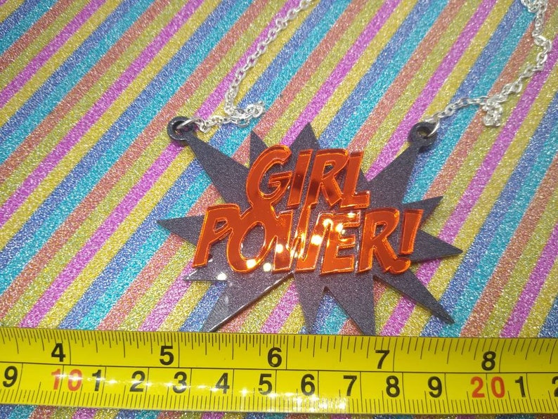 May include: A silver chain necklace with a pendant that reads "GIRL POWER!" in orange letters. The pendant is in a black starburst shape. A yellow measuring tape is at the bottom of the image.
