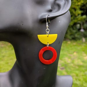 May include: A drop earring featuring a silver-toned metal hook, a yellow semi-circle shape, and a red circular ring suspended below. The earring is composed of smooth, flat geometric components connected by small silver-toned metal jump rings.