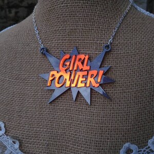 May include: A silver-toned necklace featuring a pendant with the words "GIRL POWER!" in orange, set against a gray starburst design. The necklace is displayed on a mannequin with a burlap-textured background.