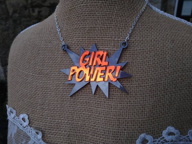 May include: A silver-toned necklace featuring a pendant with the words "GIRL POWER!" in orange, set against a gray starburst design. The necklace is displayed on a mannequin with a burlap-textured background.