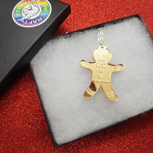 May include: A gold gingerbread man pendant necklace in a white gift box. The pendant is a mirror-like acrylic with a gingerbread man design.
