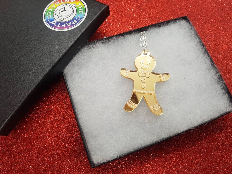 May include: A gold gingerbread man pendant necklace in a white gift box. The pendant is a mirror-like acrylic with a gingerbread man design.