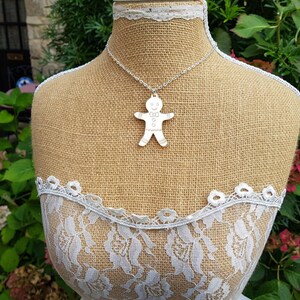 May include: A silver necklace with a gingerbread man pendant. The necklace is on a burlap mannequin with white lace trim.