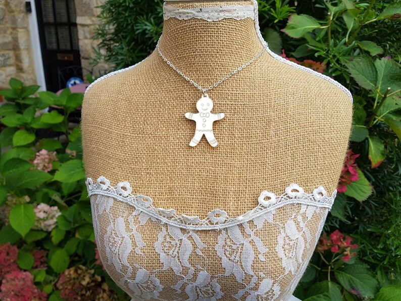 May include: A silver necklace with a gingerbread man pendant. The necklace is on a burlap mannequin with white lace trim.