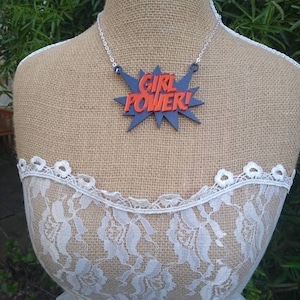 May include: A necklace with a pendant that reads "GIRL POWER!" in red letters on a blue burst design. The necklace is displayed on a mannequin wearing a white lace top and a burlap-covered torso.