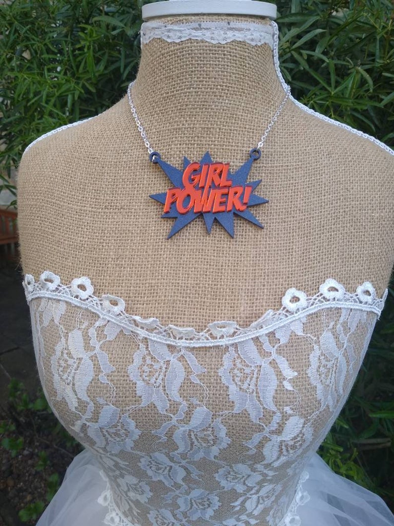 May include: A necklace with a pendant that reads "GIRL POWER!" in red letters on a blue burst design. The necklace is displayed on a mannequin wearing a white lace top and a burlap-covered torso.