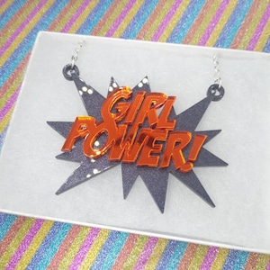 May include: A necklace with a black burst design and the words "GIRL POWER!" in orange. The necklace is displayed in a white box with a colorful background.