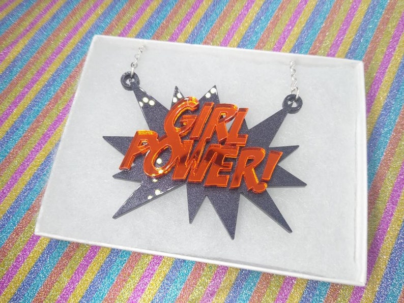 May include: A necklace with a black burst design and the words "GIRL POWER!" in orange. The necklace is displayed in a white box with a colorful background.