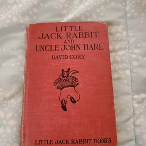 May include: A vintage red book titled "Little Jack Rabbit and Uncle John Hare" by David Cory. The cover features a black illustration of a rabbit in clothing. The book is part of the "Little Jack Rabbit Books" series.