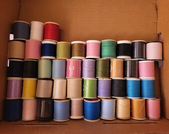 May include: A collection of colorful thread spools in a cardboard box. The spools are various colors, including red, pink, blue, green, and yellow. The threads are wound on wooden spools, perfect for sewing and crafting projects.