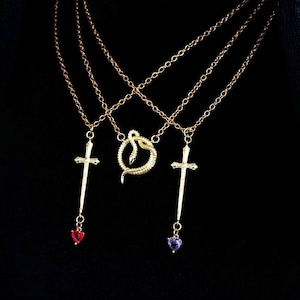 Op de afbeelding: Drie goudkleurige kettingen met hangers. Twee kettingen hebben kruisvormige hangers, een met een rode hartvormige edelsteen en de andere met een paarse hartvormige edelsteen. De derde ketting heeft een slangenhanger.