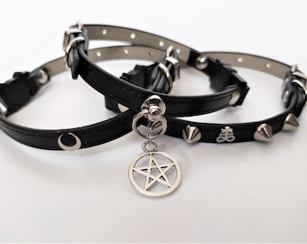 May include: Three black leather chokers with silver-tone hardware. One choker features a crescent moon charm, another has a pentagram pendant, and the third has metal spikes and a symbol. The chokers are displayed on a white surface.