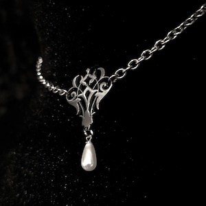 May include: A silver-colored necklace with a decorative filigree pendant and a teardrop-shaped pearl. The necklace is on a chain and is displayed against a black background. The pendant has an ornate, symmetrical design.