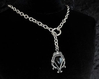 May include: Silver necklace featuring a black gemstone pendant. The pendant is diamond-shaped with intricate filigree. The necklace is displayed on a black mannequin, highlighting the chain and pendant's design.