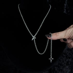 May include: A silver-toned lariat necklace featuring two four-pointed star charms. The chain connects to a central star, with a secondary chain segment extending to a second star charm, creating a layered, asymmetrical design with a metallic finish.