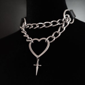 Może przedstawiać: Srebrny naszyjnik typu choker z wisiorkiem w kształcie serca i małym urokiem w kształcie sztyletu. Naszyjnik posiada czarny skórzany pasek i dwuwarstwową konstrukcję łańcucha. Biżuteria jest prezentowana na czarnym manekinie.