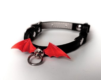 May include: A black leather cat collar with a red plastic bat wing charm. The collar has a silver metal ring for attaching a leash.