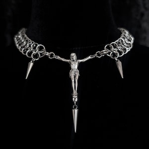 May include: Silver-toned necklace featuring a chainmail collar with three conical spikes. A detailed pendant of a crucified figure hangs from the center, with a spike pendant below. The necklace is displayed on a black surface.