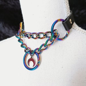 Puede incluir: Un collar gargantilla con acabado iridiscente arcoíris. El collar presenta un diseño de cadena con un anillo grande y un colgante de media luna. La gargantilla tiene una correa de cuero negro y se muestra en un maniquí blanco.