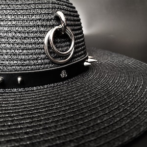 May include: Black straw hat with a wide brim, featuring a black leather band adorned with silver spikes and a decorative silver charm. A silver ring detail hangs from the hat's crown. The hat has a gothic or punk aesthetic.