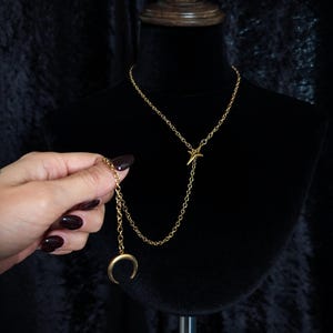 Op de afbeelding: Een goudkleurige ketting met een fijne schakelketting, een vierpuntige sterbedel in het midden en een maansikkelhanger die aan een tweede kettinggedeelte hangt, wat zorgt voor een gelaagd Y-vormig ontwerp op een zwarte displayhouder.
