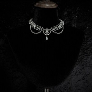 K&ouml;nnte beinhalten: Ein silberfarbenes Choker-Halsband mit Kettengeflecht-Design. Die Halskette hat einen zentralen Anh&auml;nger mit herzf&ouml;rmigem Element und eine herabh&auml;ngende tropfenf&ouml;rmige Perle. Die Halskette wird auf einer schwarzen Schaufensterpuppe pr&auml;sentiert.