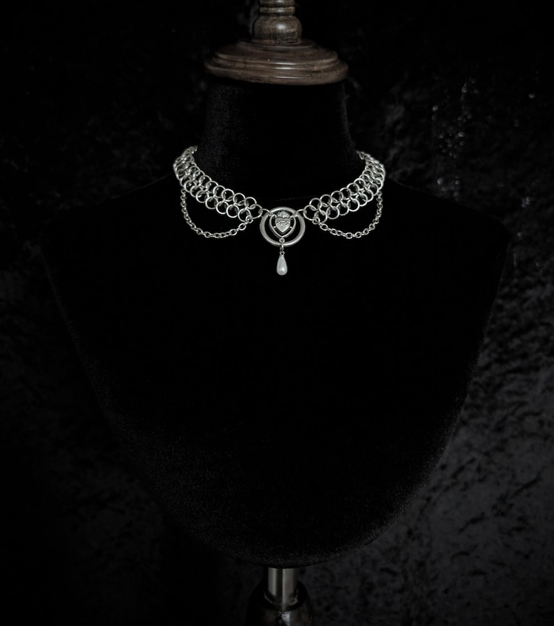 K&ouml;nnte beinhalten: Ein silberfarbenes Choker-Halsband mit Kettengeflecht-Design. Die Halskette hat einen zentralen Anh&auml;nger mit herzf&ouml;rmigem Element und eine herabh&auml;ngende tropfenf&ouml;rmige Perle. Die Halskette wird auf einer schwarzen Schaufensterpuppe pr&auml;sentiert.