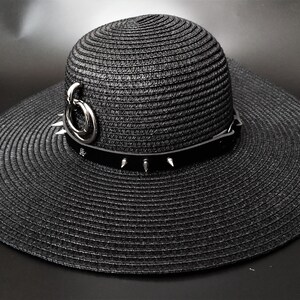 May include: Black wide-brimmed sun hat with a decorative band featuring silver spikes and a silver ring accent. The hat is made of woven material and has a structured crown and a wide, floppy brim. A stylish accessory.