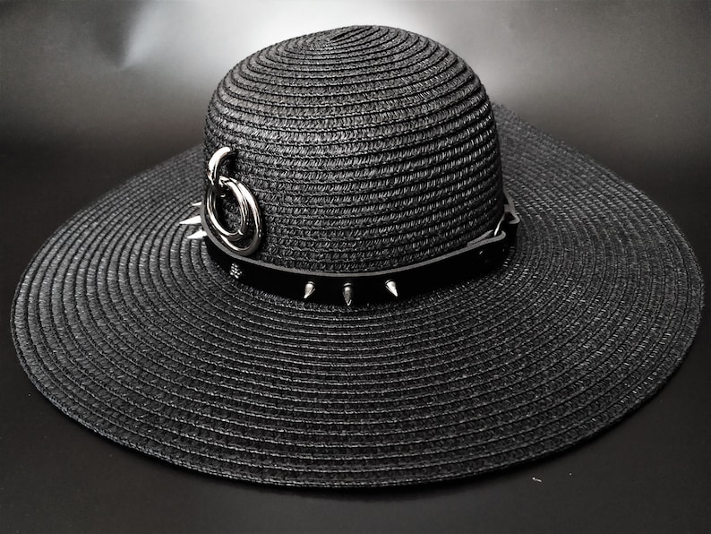 May include: Black wide-brimmed sun hat with a decorative band featuring silver spikes and a silver ring accent. The hat is made of woven material and has a structured crown and a wide, floppy brim. A stylish accessory.