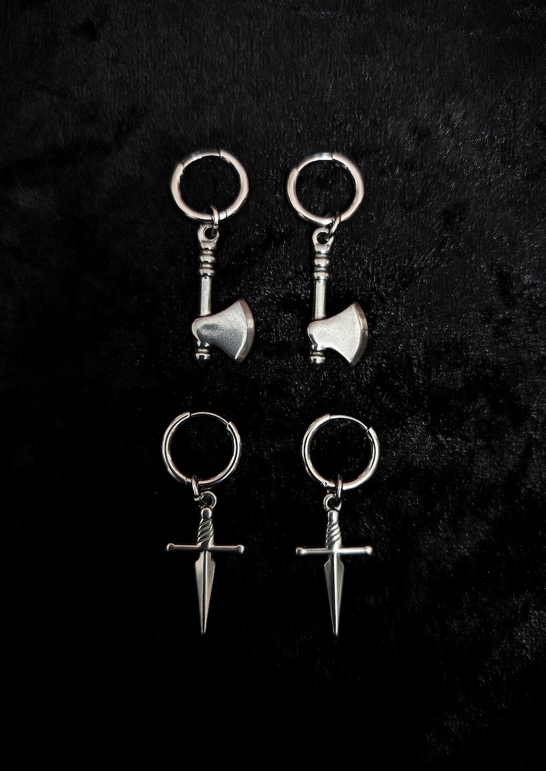 May include: Four silver-toned earrings on a black background. Two pairs of earrings are visible: one pair features small axe pendants, and the other pair has dagger pendants. Each earring has a circular hoop for attachment.