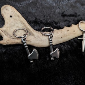 May include: Three pairs of silver-toned metal earrings. Two pairs feature axe pendants, and the third pair has sword pendants. The earrings are displayed on a bone against a dark, textured background.