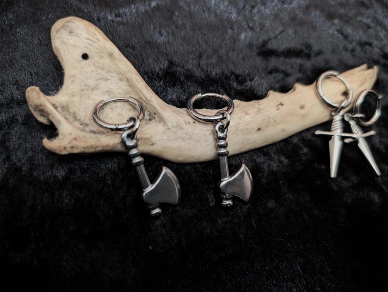 May include: Three pairs of silver-toned metal earrings. Two pairs feature axe pendants, and the third pair has sword pendants. The earrings are displayed on a bone against a dark, textured background.