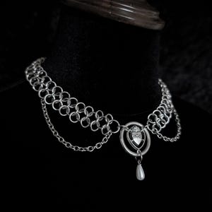 K&ouml;nnte beinhalten: Silberne Kettenhemd-Halskette mit Herzanh&auml;nger und h&auml;ngender Perle. Die Halskette wird auf einer schwarzen Oberfl&auml;che pr&auml;sentiert. Der Choker hat mehrere Reihen aus Kettenhemd und ein filigranes Design.