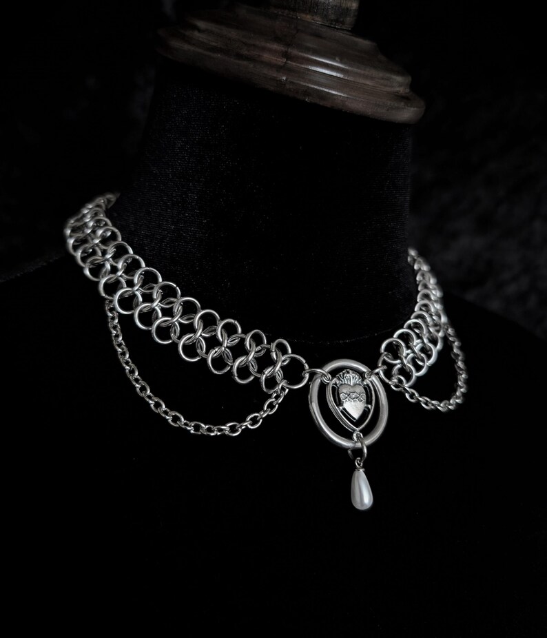 K&ouml;nnte beinhalten: Silberne Kettenhemd-Halskette mit Herzanh&auml;nger und h&auml;ngender Perle. Die Halskette wird auf einer schwarzen Oberfl&auml;che pr&auml;sentiert. Der Choker hat mehrere Reihen aus Kettenhemd und ein filigranes Design.