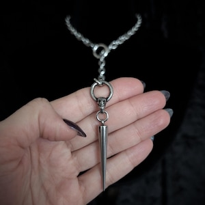 May include: A silver-toned necklace with a chain and a pendant featuring a long, pointed spike. The pendant is attached to a ring with a small skull charm. The necklace is displayed against a dark background.