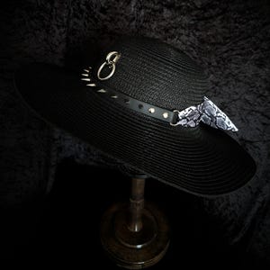 May include: A black wide-brimmed hat featuring a textured straw-like finish. The hat is adorned with a black band decorated with silver-toned metal spikes, a circular ring hardware piece, and a patterned fabric scarf with a grey and white snakeskin print.
