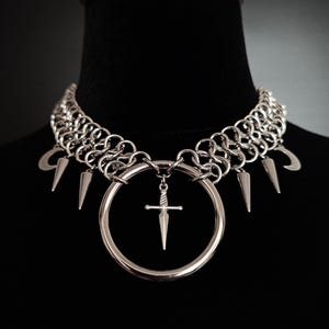 May include: Silver-toned choker necklace with a chainmail design. The necklace features a large circular pendant with a small sword charm and several spike and crescent moon charms. The jewelry is displayed on a black mannequin.