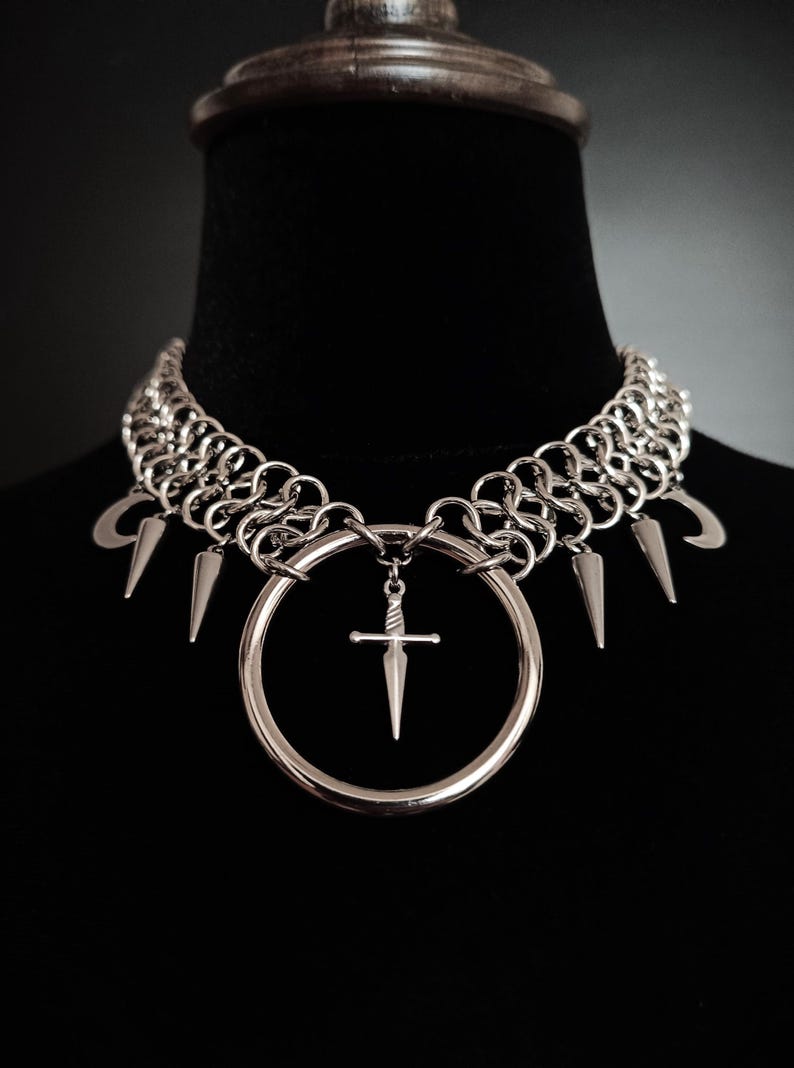 May include: Silver-toned choker necklace with a chainmail design. The necklace features a large circular pendant with a small sword charm and several spike and crescent moon charms. The jewelry is displayed on a black mannequin.