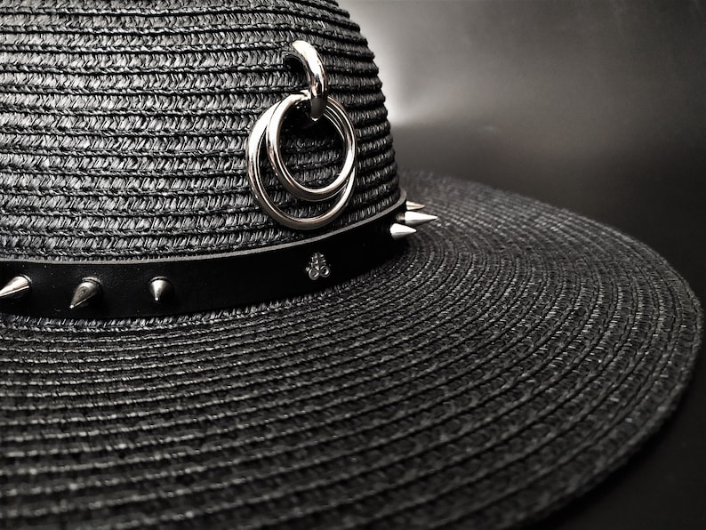 May include: Black straw hat with a wide brim, featuring a black leather band adorned with silver spikes and a decorative silver charm. A silver ring detail hangs from the hat's crown. The hat has a gothic or punk aesthetic.