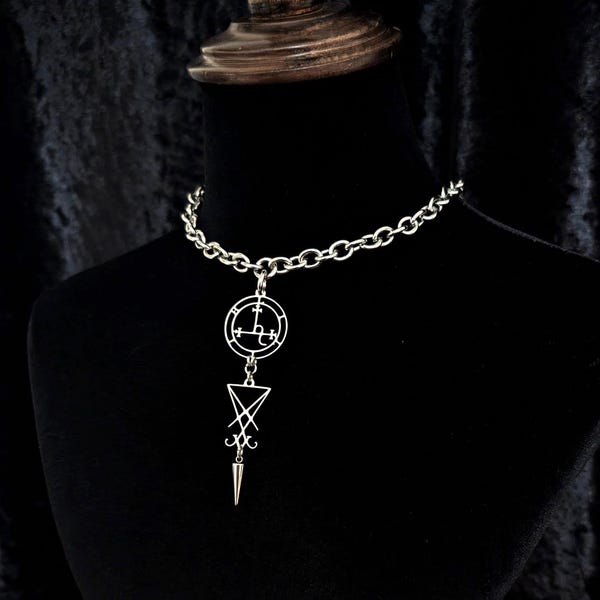 May include: Silver-toned necklace with a large chain and a pendant featuring a circular design with symbols and a downward-pointing triangle with a spike. The necklace is displayed on a black mannequin.