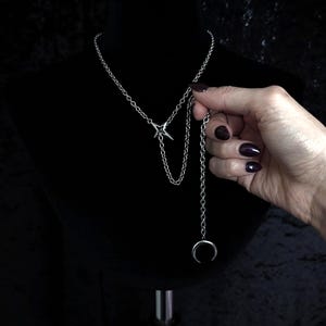 May include: Silver-toned necklace with a celestial theme. The necklace features a chain with a star-shaped pendant and a dangling chain ending in a crescent moon. The jewelry is displayed on a black mannequin.