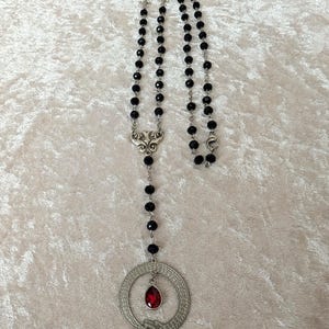 May include: A black beaded necklace with a silver pendant featuring a snake coiled around a red gemstone. The pendant is in the shape of a circle.
