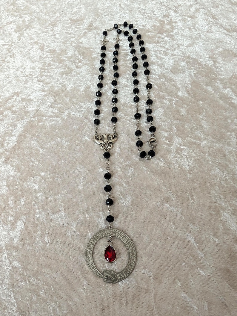 May include: A black beaded necklace with a silver pendant featuring a snake coiled around a red gemstone. The pendant is in the shape of a circle.