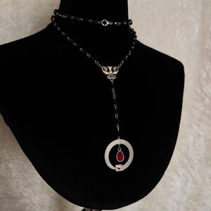 May include: A silver necklace with a black beaded chain and a pendant featuring a snake coiled around a circle with a red gemstone in the center.