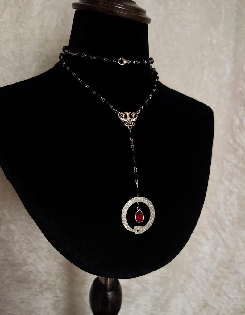 May include: A silver necklace with a black beaded chain and a pendant featuring a snake coiled around a circle with a red gemstone in the center.