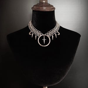 May include: Silver chainmail choker necklace with a large circular pendant featuring a cross design. The necklace also includes small crescent moon and spike charms. The jewelry is displayed on a black mannequin.