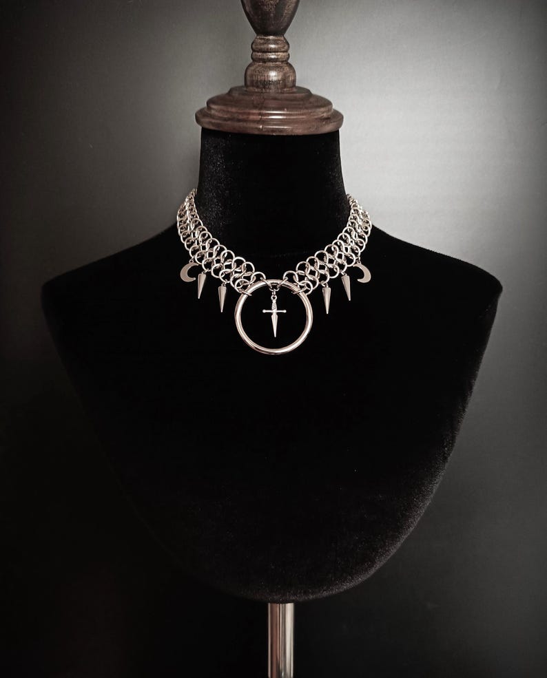 May include: Silver chainmail choker necklace with a large circular pendant featuring a cross design. The necklace also includes small crescent moon and spike charms. The jewelry is displayed on a black mannequin.