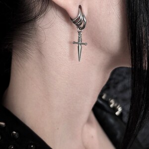 May include: A silver-colored earring with a small dagger pendant. The earring is a hoop style with multiple rings, and the dagger hangs from the bottom. The image is a close-up, showing the earring against a blurred background.