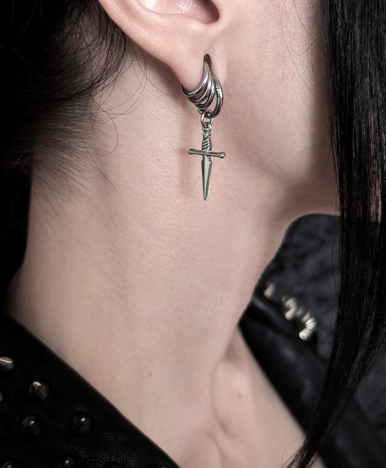 May include: A silver-colored earring with a small dagger pendant. The earring is a hoop style with multiple rings, and the dagger hangs from the bottom. The image is a close-up, showing the earring against a blurred background.